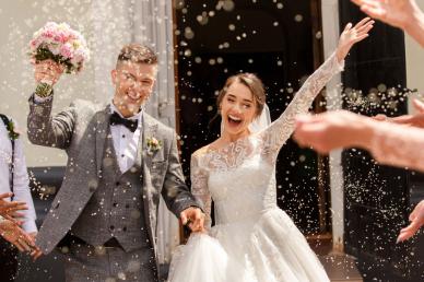 A joyful bride and groom walk out of a building as guests throw confetti, celebrating their wedding day.