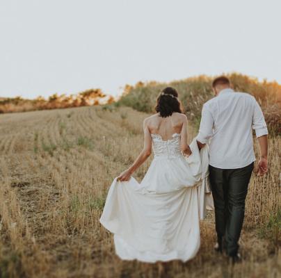 A bride and groom walk hand in hand through a golden field at sunset, with the bride lifting her dress slightly off the ground.