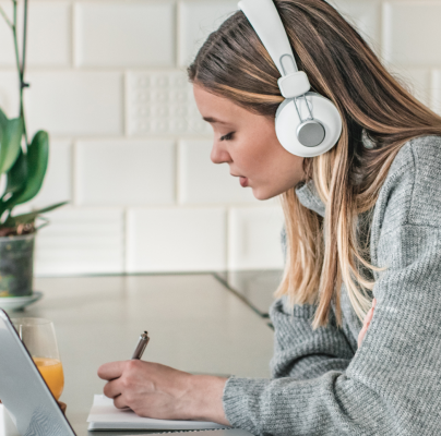 Woman wearing headphones writing in notebook while using laptop