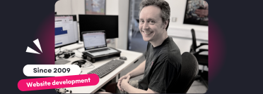A black-and-white photo of a website development specialist smiling while working at a desk with multiple monitors displaying code and documents